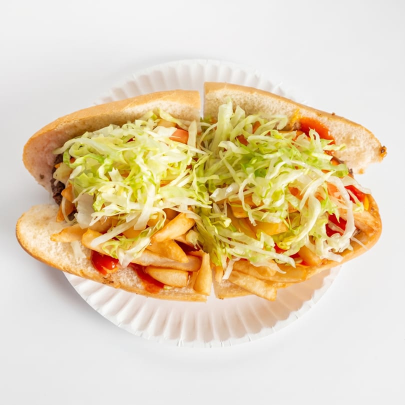 A large sub sandwich, cut in half, filled with fries, shredded lettuce, and ketchup on a paper plate.